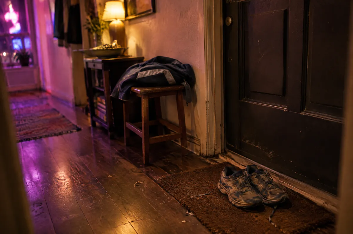 Flur am Abend neben der Wohnungstür mit Laufschuhen auf der Fußmatte und einer gefalteten Jacke auf einem Hocker
